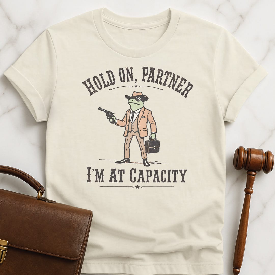funny new attorney graphic tee that says Hold on partner Im at capacity featuring cowboy lawyer toad or frog holding a gun and briefcase in cream