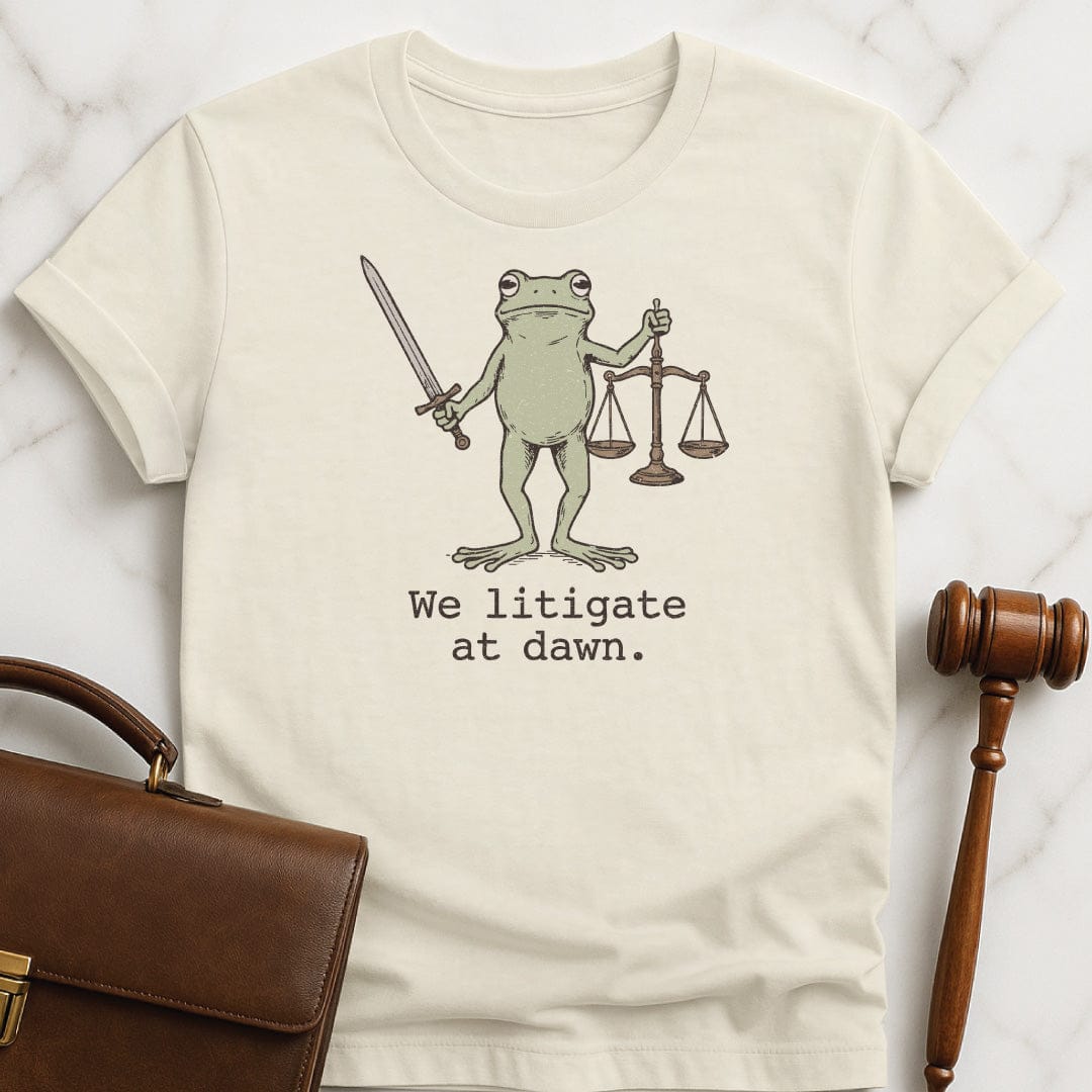 funny attorney t shirt that says We litigate at dawn featuring frog with a sword and scales of justice in cream
